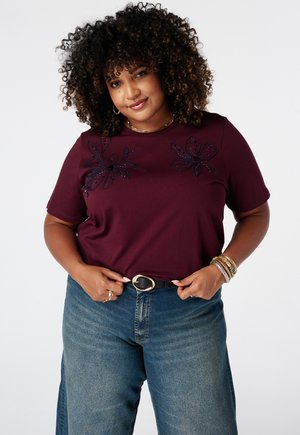 Femme aux cheveux bouclés portant une chemise bordeaux à motifs floraux et un jean bleu, ajustant une ceinture noire avec une boucle dorée.