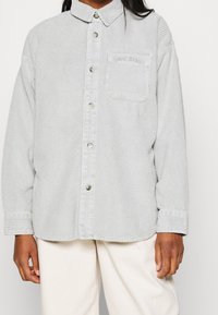 Osoba nosząca jasnoszary, kordowy, zapinany na guziki shirt z kieszenią na piersi oraz kremowe spodnie, pokazana od ramion do połowy uda.