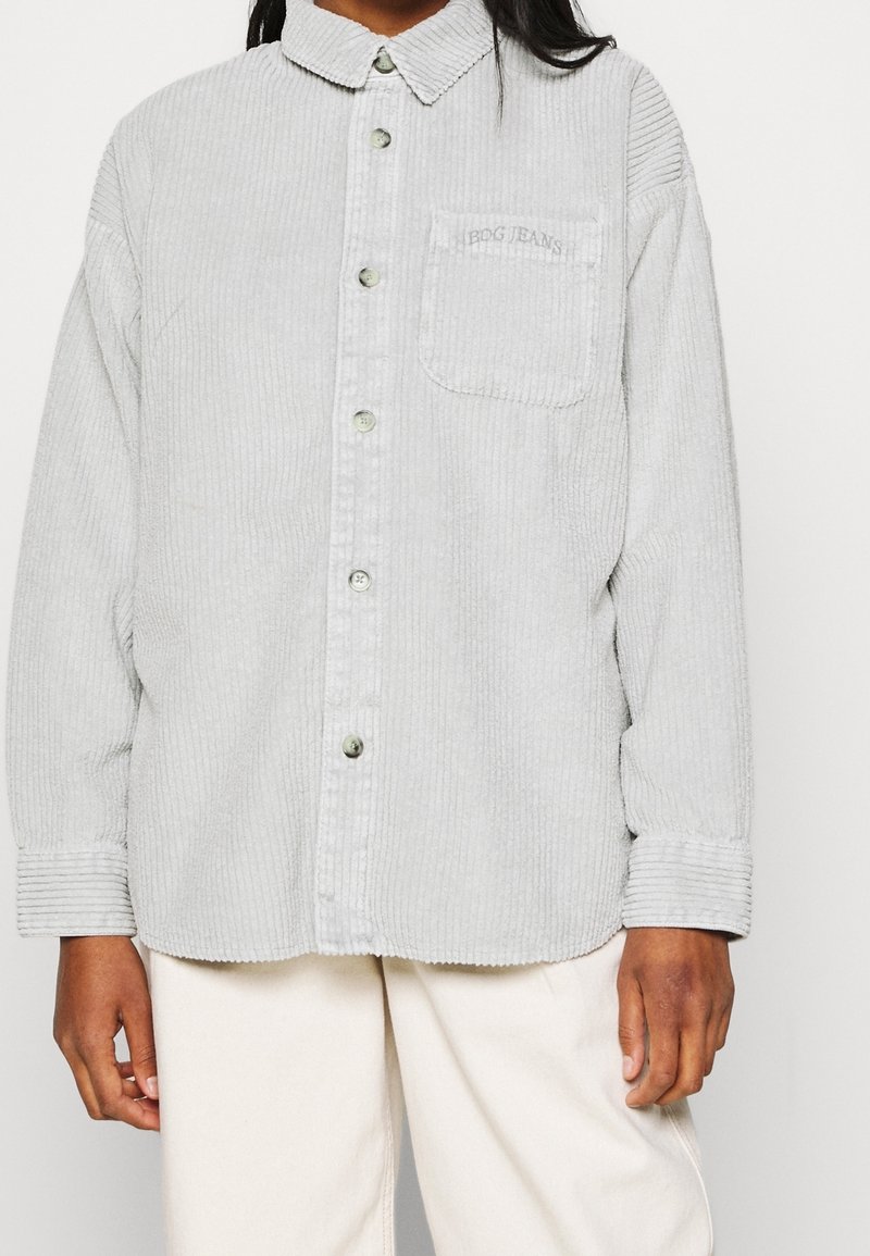 Osoba nosząca jasnoszary, kordowy, zapinany na guziki shirt z kieszenią na piersi oraz kremowe spodnie, pokazana od ramion do połowy uda.