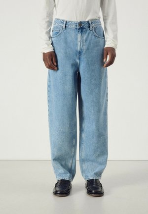 Personne portant un jean bleu clair à jambes larges, des chaussures en cuir noires et un haut blanc à manches longues, debout devant un fond clair uni.