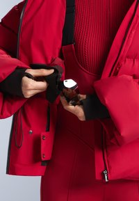 Piros, fekete részletekkel és cipzárral ellátott puffertalpú dzseki. Egy személy egy zsebben fényvisszaverő tárgyat tart, amely felfedi a fekete, texturált belsőt.