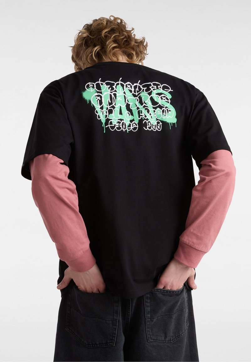 Personne avec des cheveux bouclés portant un t-shirt noir par-dessus des manches longues roses, avec un texte style graffiti vert et blanc dans le dos, les mains dans les poches.