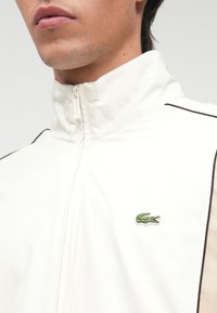 Λευκό μπουφάν με φερμουάρ και ψηλό γιακά, με μαύρες λεπτομέρειες και ένα μικρό πράσινο λογότυπο κροκόδειλου στο αριστερό στήθος.