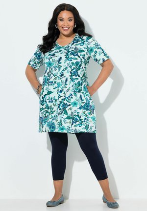 Frau lächelt mit Händen in den Taschen, trägt eine blau-grüne, floral gemusterte Tunika, schwarze Leggings und blaue Ballerinas vor weißem Hintergrund.