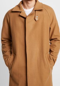 Homme portant un manteau en laine couleur camel avec un col et un rabat à boutons-pression, mains dans les poches latérales, sur un t-shirt blanc à col rond.