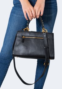 Personne portant un jean bleu tenant un sac à main en cuir noir avec une fermeture éclair dorée et une bandoulière amovible.