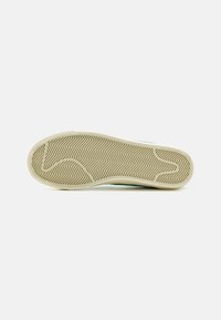 Semelle de chaussure avec une bande de roulement en caoutchouc beige en motif chevrons et rainures courbes sur fond blanc.
