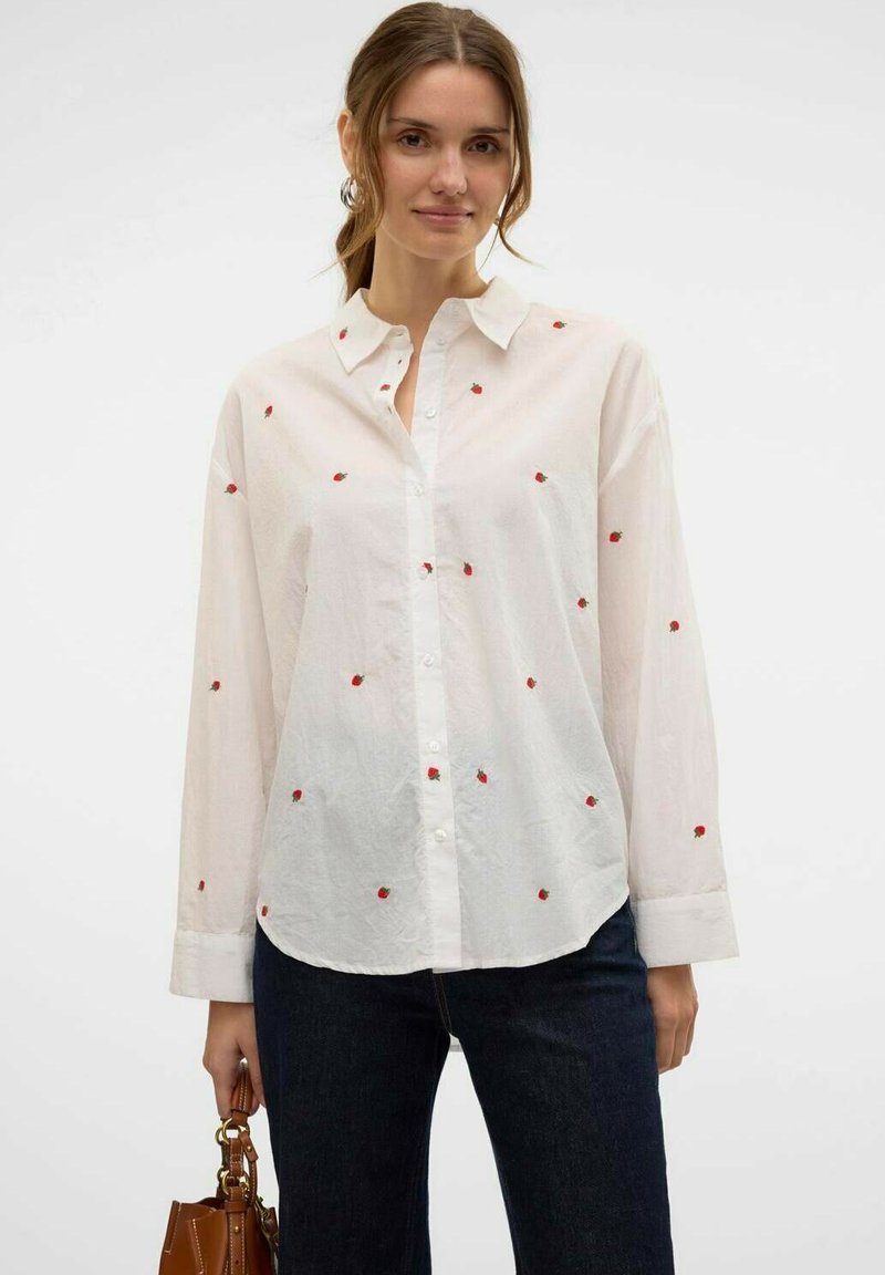 Camicia bianca con dettagli di fragola ricamati in rosso, colletto button-down, maniche lunghe, vestibilità leggermente ampia, indossata con jeans scuri e una borsa.
