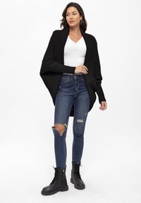 Cardigan nero a coste con apertura frontale, indossato sopra una maglietta bianca aderente. Jeans strappati blu scuro e stivali militari neri stringati completano il look.