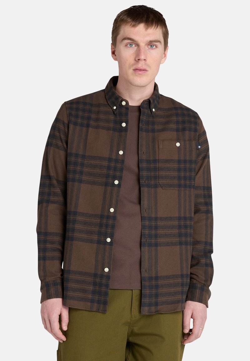 Camicia a quadri marrone con linee blu scuro, caratterizzata da un colletto button-down, taschino sul petto e bottoni in plastica, indossata sopra una maglietta marrone.