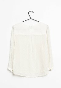 Blusa bianca a maniche lunghe con motivo a pois in rilievo, scollatura rotonda e polsini con bottoni. Realizzata in tessuto leggero e morbido. Appesa a un gancio.