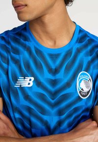 T-shirt de sport bleu avec un motif de vague en noir, un col rond et un logo blanc sur le côté gauche de la poitrine avec un emblème circulaire.