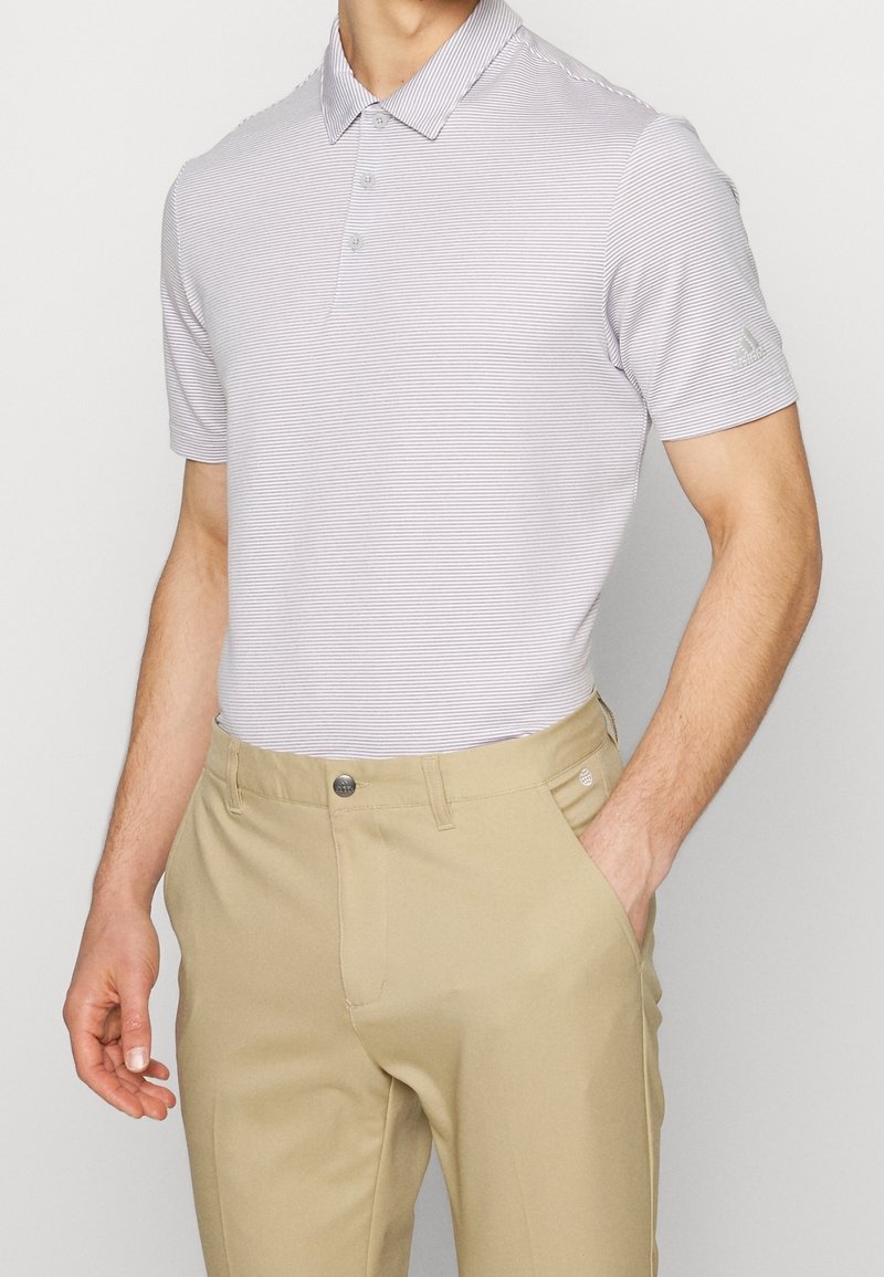 Homme portant un polo à manches courtes rayé gris clair et un pantalon beige, une main dans la poche, sur fond uni.