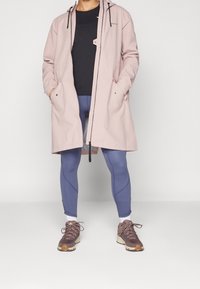 Veste imperméable rose clair avec capuche à cordon noir, associée à des leggings bleu marine et des baskets marron avec des accents blancs et orange.