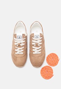 Ζευγάρι από μπεζ σουέντ αθλητικά παπούτσια με λευκά κορδόνια, ετικέτες λογότυπου στη γλώσσα, και δύο περιτυλιγμένα πορτοκαλί κορδόνια τοποθετημένα δίπλα τους σε λευκό φόντο.