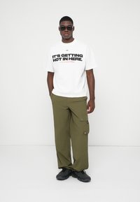 Homem com óculos de sol pretos, t-shirt branca com o texto "ESTÁ A FICAR QUENTE AQUI", calças cargo olivas e sapatos pretos, de pé contra um fundo branco.