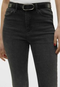 Jeans de denim negro con un ajuste ajustado, que cuentan con un diseño estándar de cinco bolsillos y un cinturón negro brillante con una hebilla ovalada de plata.