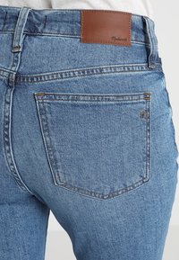 Blue denim jeans with a smooth texture, featuring a brown leather patch at the waistband and two back pockets with subtle stitching details.