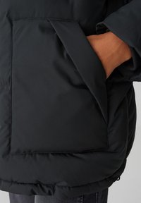 Giacca nera imbottita con una grande tasca frontale e una texture liscia e opaca. Il tessuto è leggermente lucido, con una vestibilità rilassata all'orlo.