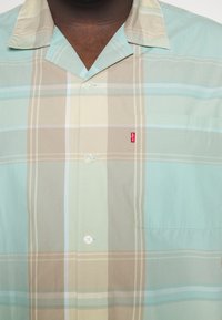 Camicetta a maniche corte a quadri in verde chiaro, beige e crema. Presenta un design con bottoni, tasca sul petto e etichetta rossa del marchio sulla tasca.