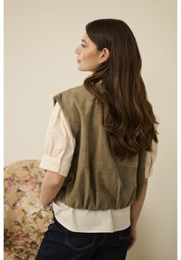 Olivengrøn ruskindsvest med en rynket kant, lagdelt over en cremefarvet bluse. Bæres med mørke jeans, der har korte pufærmer og knapper.