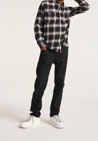 Camicia a maniche lunghe a scacchi bianco e nero con due tasche sul petto, abbinata a pantaloni neri e sneaker bianche con accenti argentati.