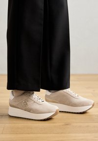 Baskets en daim beige avec une semelle plateforme blanche, présentant un design à lacets, un logo en relief et des accents texturés. Associées à un pantalon noir.