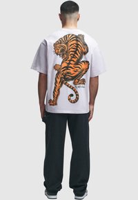 Hvit oversized t-skjorte med et oransje og svart tiger-graphic på ryggen. Svart bukser og hvite joggesko fullfører antrekket.
