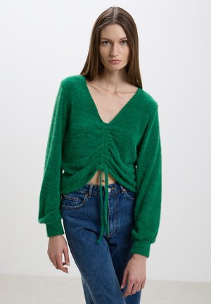 Maglione verde a trama con scollatura a V e dettaglio con cordoncino in vita; maniche lunghe e ampie; abbinato a jeans blu a vita alta.