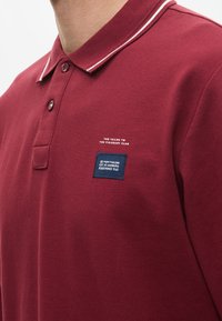 Bordeaux polo shirt van een gestructureerde stof, met een knoopsluiting met twee knopen en contrasterende witte kraagafwerking. Voorzien van logopatches op de borst.