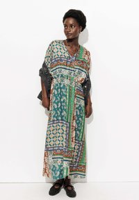 Femme aux cheveux courts et bouclés portant une robe maxi en patchwork multicolore, tenant une veste en denim et des ballerines noires à clous.
