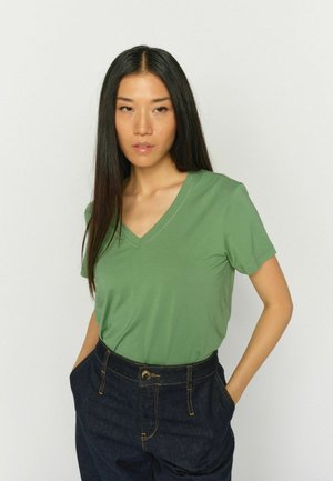 Žena s dlouhými tmavými vlasy, která má na sobě zelené tričko s V-výstřihem a tmavě modré džíny, stojí s rukama v kapsách před jednobarevným pozadím.