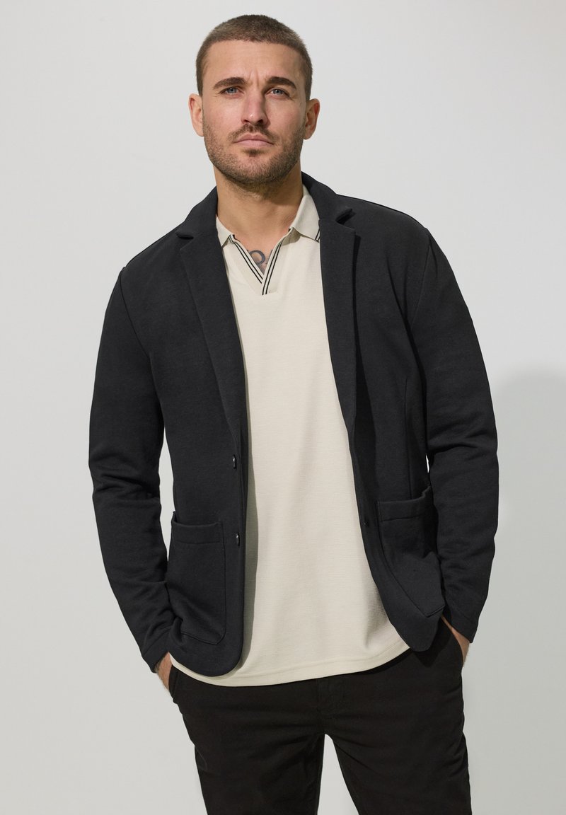 Hombre con cabello corto y barba, vestido con blazer negro sobre camisa polo beige claro, de pie con las manos en los bolsillos contra un fondo liso.
