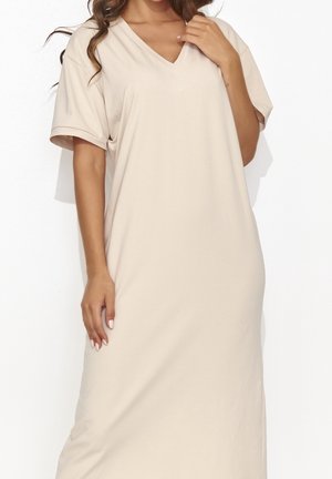Vrouw draagt een losse beige knielange jurk met korte mouwen en een V-hals, staand met één hand bij haar sleutelbeen.
