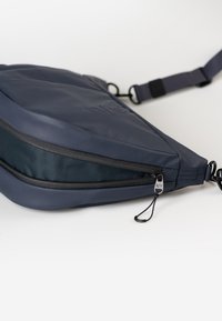 Borsa a tracolla triangolare blu navale con superficie testurizzata, cerniera nera e tracolla regolabile. Dettagli notevoli includono un logo e un piccolo tirante in metallo per la cerniera.
