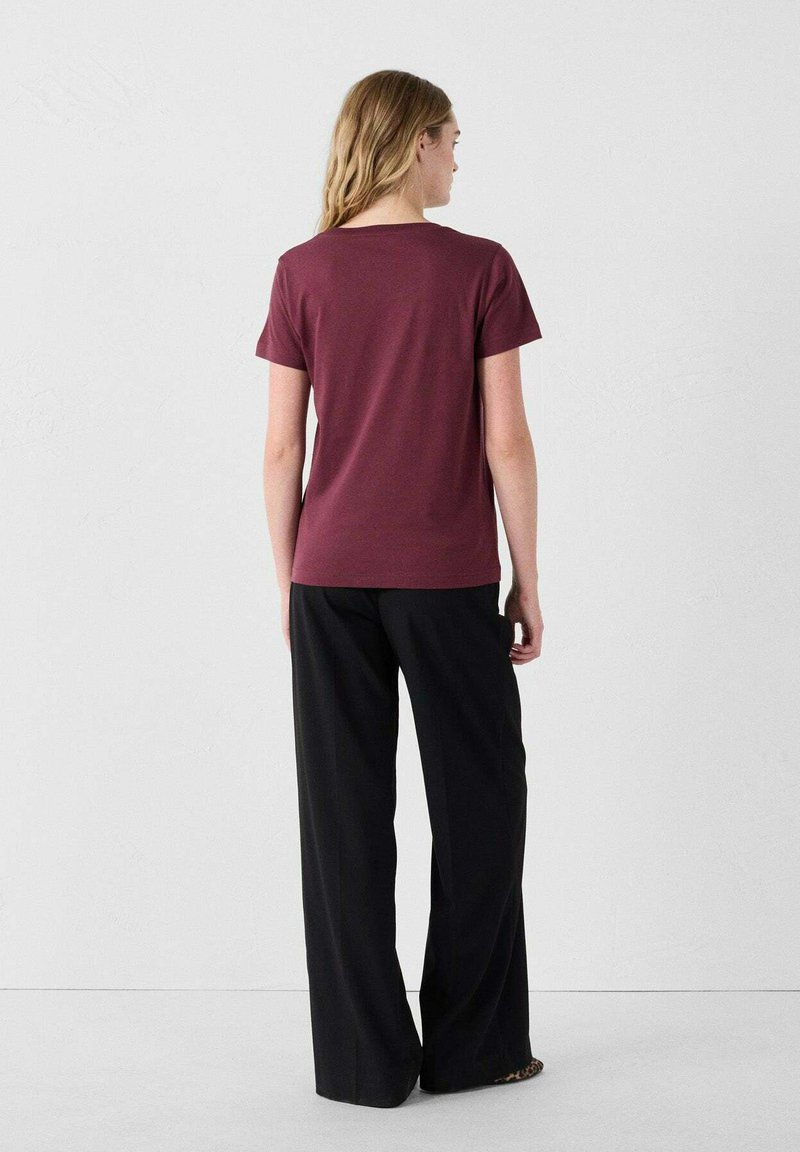 Camiseta burdeos de manga corta con cuello redondo, combinada con pantalones negros holgados, vista desde la parte trasera contra un fondo liso.
