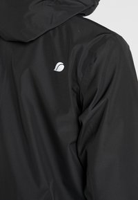 Veste imperméable noire avec capuche et un discret logo blanc sur l'épaule. Le tissu présente une texture lisse et mate.