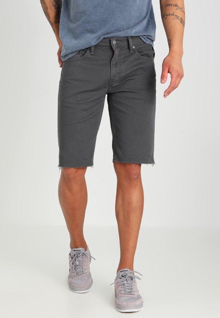 Homme portant un short en jean gris foncé avec des ourlets bruts, des baskets grises Reebok et un t-shirt bleu, debout devant un fond clair uni.