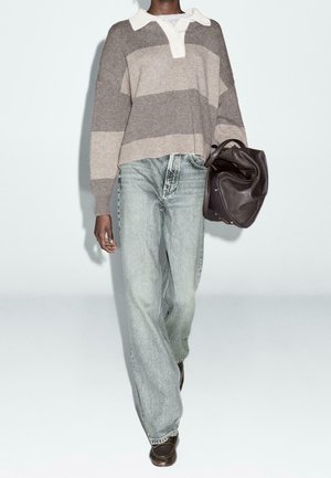 Person trägt einen grau-beigen gestreiften Pullover, hellblaue Jeans, dunkle Schuhe und trägt eine große dunkelbraune Ledertasche.