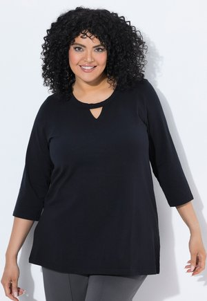 Femme aux cheveux bouclés souriante, portant un haut noir à manches trois-quarts et un pantalon gris sur fond blanc.