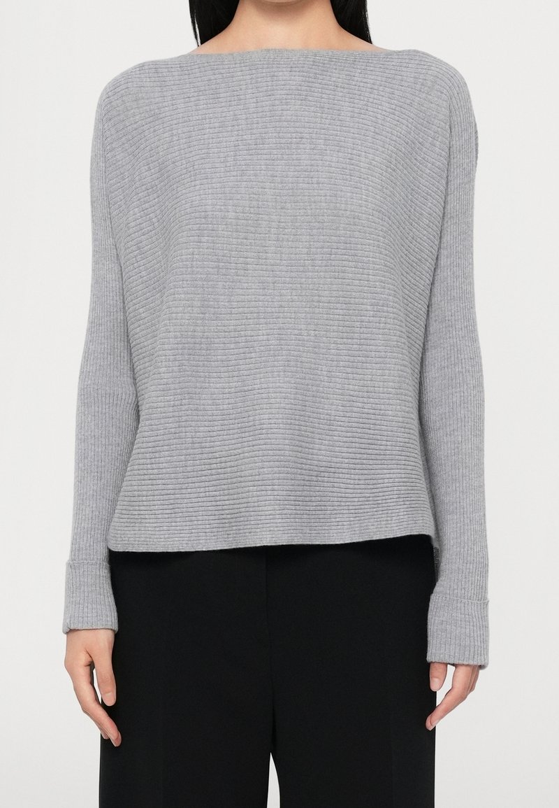 Femme portant un pull en maille côtelée gris clair à manches longues et un pantalon noir à jambes larges, debout devant un fond uni.