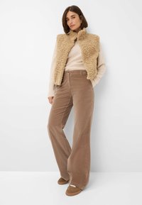 Beige nep-bont vest over een crèmekleurige trui, gecombineerd met lichtbruine fluwelen flared broek en bruine suède instappers. Eenvoudig ontwerp.
