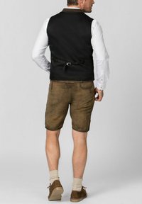 Schwarze Weste mit verstellbarem Rückengurt, kombiniert mit braunen Shorts. Braune Lederschuhe und helle Socken runden das Outfit ab.