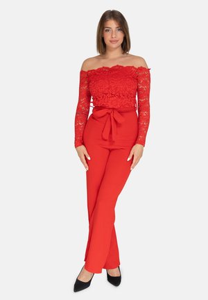 Mujer de pie con un top de encaje rojo con los hombros descubiertos y pantalones rojos de talle alto con lazo delantero, combinados con tacones negros puntiagudos.
