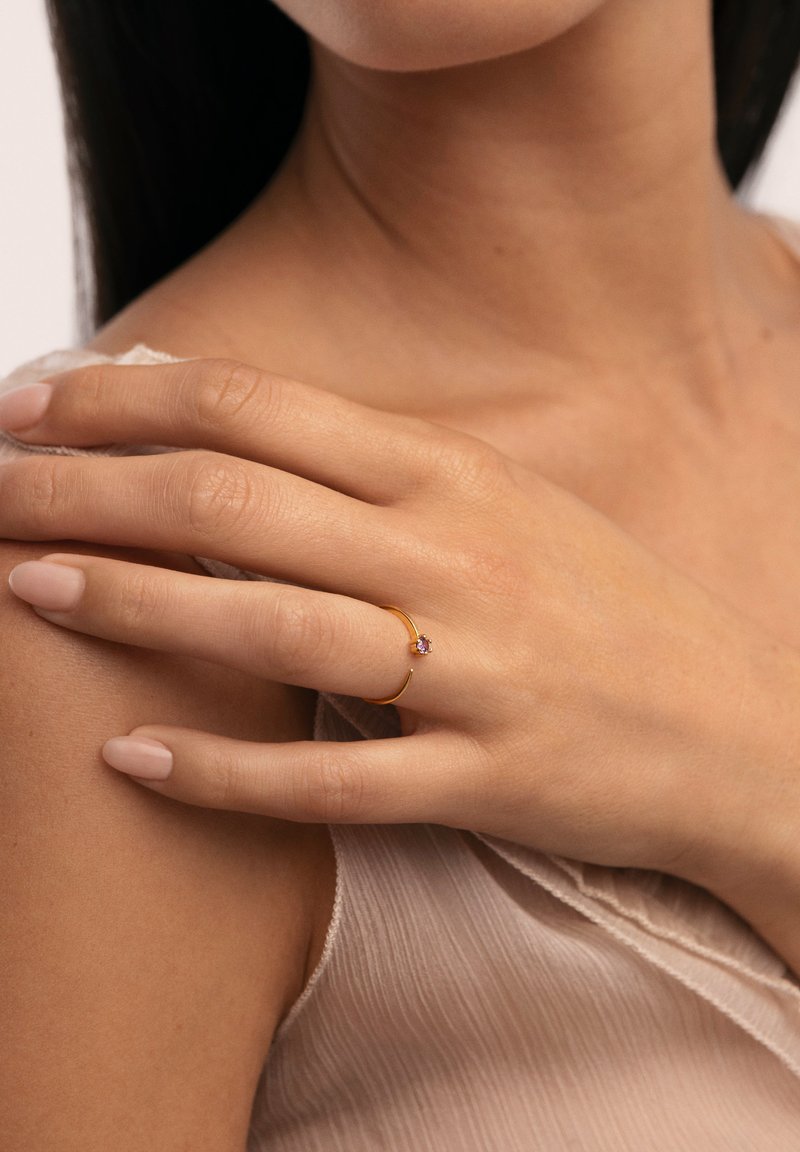 Anillo de oro con una banda delgada y un pequeño engaste de gema púrpura, llevado en una mano con uñas bien cuidadas sobre un fondo suave y texturizado.