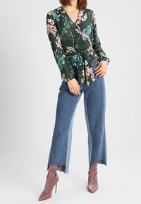 Grön blommig wrap-top med långa ärmar och knytning i midjan, parat med vida blå denimjeans och ljusrosa högklackade stövlar.