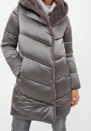 Kobieta ubrana w długi, błyszczący szary puchowy płaszcz z dużym kołnierzem obszytym futrem, z rękami w kieszeniach, na jasnoszarych spodniach.