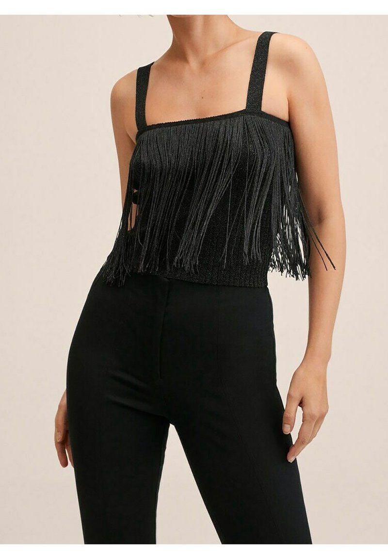 Femme portant un crop top noir à franges avec de larges bretelles et un pantalon noir taille haute sur un fond beige.