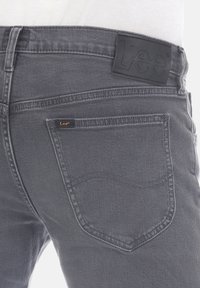 Grijze denim jeans met een gestructureerde afwerking, voorzien van een zwart leren patch met het "Lee" logo en een klein oranje label op de achterzak.