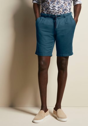 Türkise Baumwollmischung-Shorts mit Seitentaschen, umgeschlagenen Säumen und einem Kordelzugbund, kombiniert mit beigen Slippern. Der Hintergrund ist neutral.